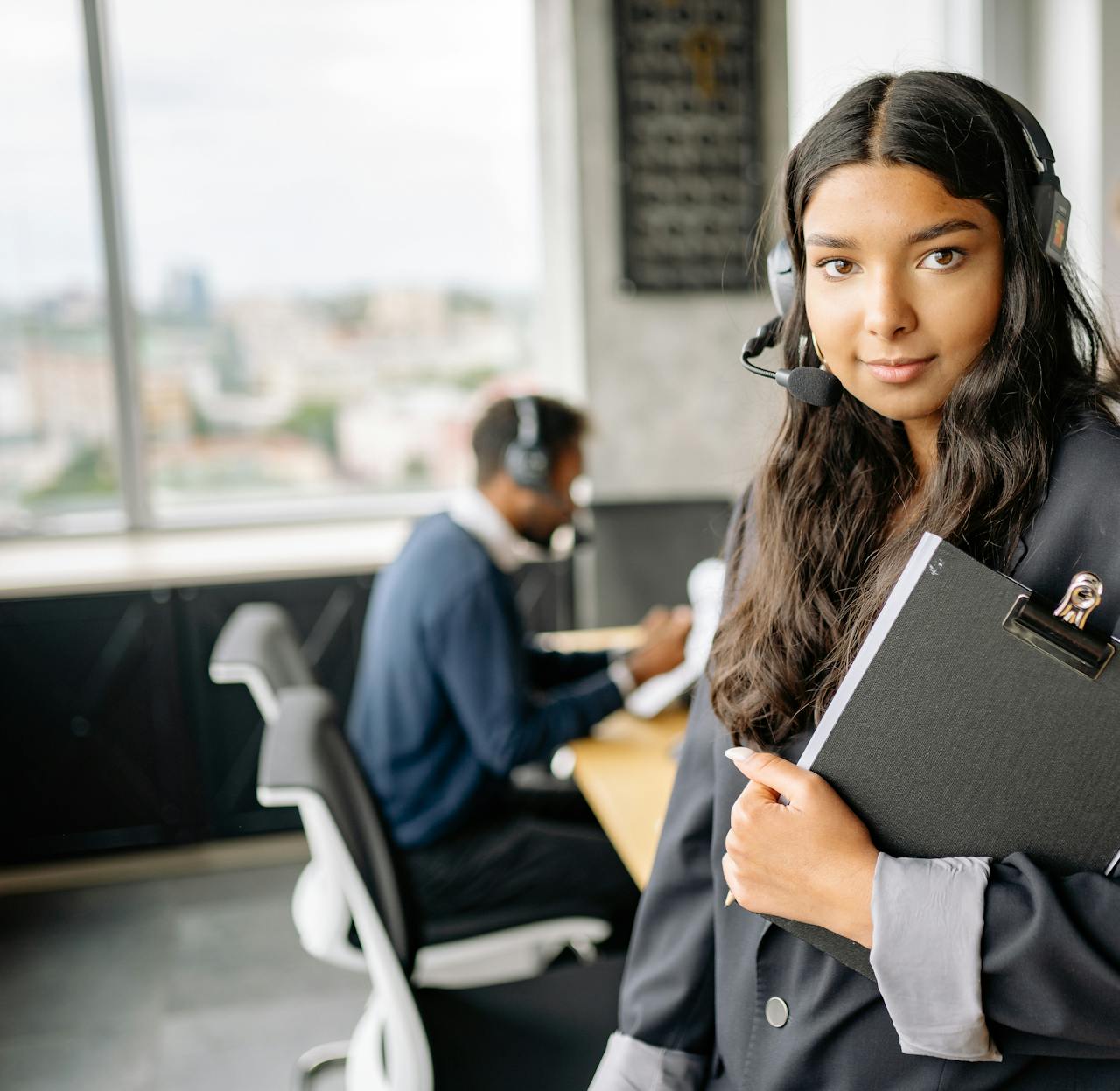 Confident woman wearing a headset and holding a clipboard in a busy office setting.